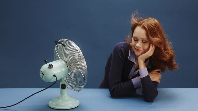 A young white ginger woman on a blue background being hot and turning on the fan 