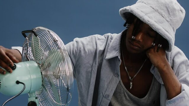 A black young man on a blue background being hot and turning on the fan 