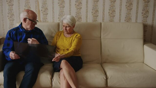 Concerned and confused old caucasian grey-haired couple looking at lungs x-ray results and worrying about diagnosis while sitting on a beige leather sofa at home together. High quality 4k footage