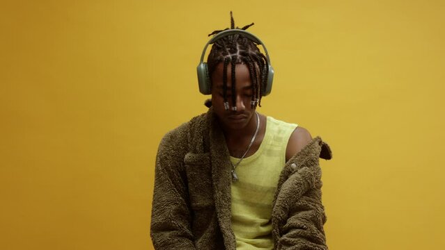 A young stylish black man, wearing a vest and white trousers, sitting on a chair enjoying some music on his headphones, in front of a yellow background - Powered by Adobe