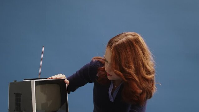 A young ginger white woman attempting to switch on an old broken retro television on, wearing a purple dress in front of a blue background sitting at a blue desk