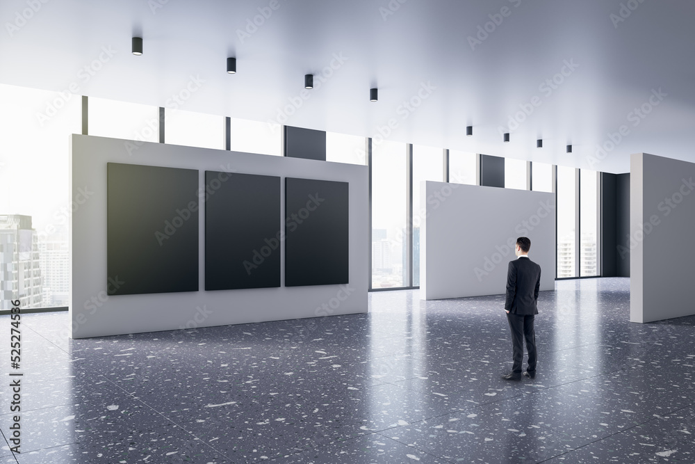 Poster back view of young man thinking in modern bright gallery interior with mock up posters and windows w