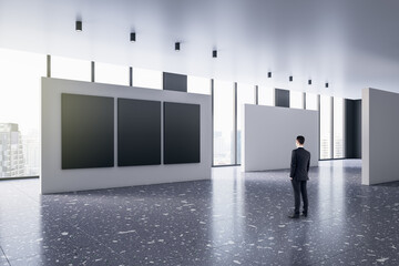 Fototapeta premium Back view of young man thinking in modern bright gallery interior with mock up posters and windows with city view.