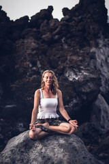 Naklejka na ściany i meble beautiful woman sitting in meditation against dark rocks