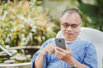 Fototapeta premium An old man is using a smartphone on a chair in the garden. Social network concept.