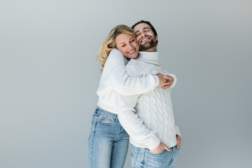 cheerful woman hugging man in white sweater posing with hands in pockets isolated on grey