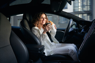 Attractive young woman holding cup of coffee and croissant while sitting on driver seat in electric car with vehicle autopilot system. Cheerful businesswoman having breakfast in electric vehicle.