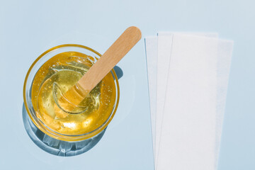 Liquid yellow sugar paste, wooden spatula, depilatory strips on a blue background. Removing unwanted hair. Sugaring. Depilation. Epilation. Beauty. Top view. - obrazy, fototapety, plakaty