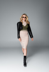 full length of blonde woman in black leather jacket and stylish sunglasses walking on grey
