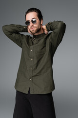 bearded man in stylish green shirt and sunglasses looking at camera while posing isolated on grey