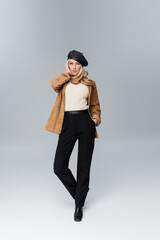 blonde woman in stylish leather beret and beige blazer posing with hand in pocket on grey