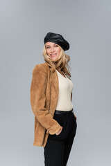 Fototapeta premium happy blonde woman in stylish leather beret and beige blazer posing with hands in pockets isolated on grey