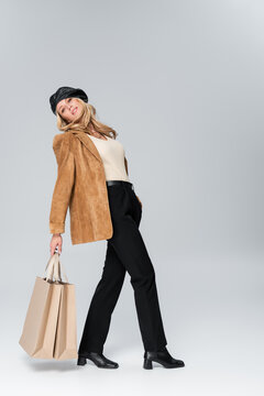 full length of blonde woman in stylish leather beret and beige blazer posing with shopping bags on grey