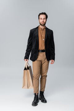 full length of bearded man in stylish autumnal outfit posing with shopping bags on grey