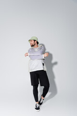 full length of bearded man in trendy sportive outfit and panama hat posing on grey