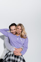 happy man hugging cheerful woman in purple sweatshirt while looking at camera on grey