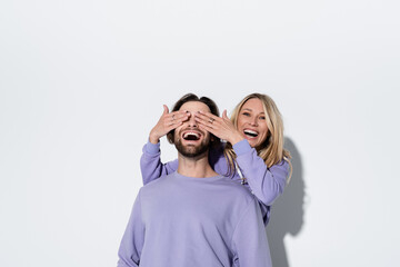 happy blonde woman covering eyes of bearded man in purple sweatshirt on grey