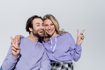 happy couple in purple sweatshirts showing peace sign and smiling isolated on grey