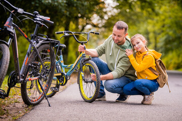 Dad and daughter inspect wheel of children's teenage bicycle on the autumn path of the park.