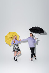 full length of joyful couple in purple sweatshirts holding hands while standing under umbrellas on grey