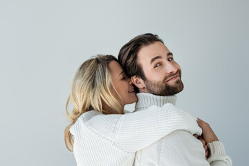 cheerful blonde woman in white knitted sweater hugging bearded man isolated on grey