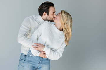 bearded man and blonde woman in knitted sweaters kissing isolated on grey