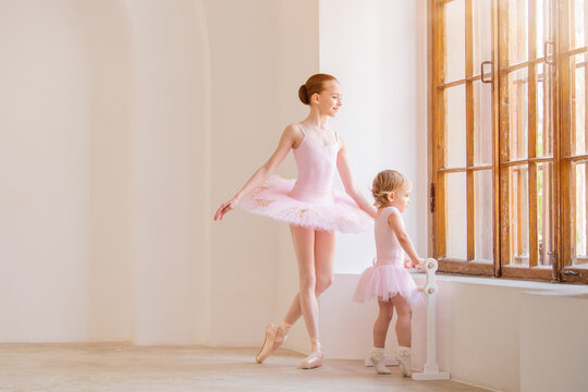 The Older Sister, A Ballerina In A Pink Tutu And Pointe Shoes, Shows The Baby How To Practice .