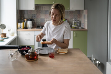Middle-aged unkempt woman spreads jam on toast having breakfast alone. Mature blonde female eats healthy food in morning thinking about own lifestyle. Lady prepares humble breakfast in late morning - Powered by Adobe