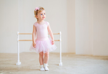 A cute blue-eyed baby ballerina in a pink leotard and tutu standing near a ballet barre. Dream...