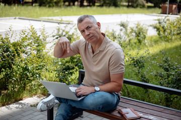 Elderly man sitting on wooden bench with crossed leg in park in summer, holding open laptop,...