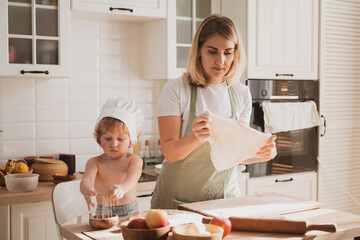 young woman in chef's apron and her little son are preparing apple pie at home in cozy kitchen
