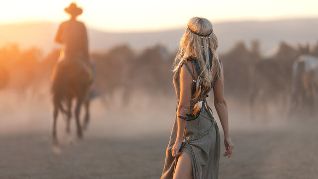 Blonde woman in traditional Indian western dress on the background of a herd of horses and a cowboy in the rays of sunset. 16x9 cinematic picture.