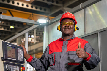 Cheerful African American CNC Machine Operator Standing Next To CNC Controller Console And Giving Thumb Up. Portrait Of Black Machine Operator In Red Helmet, Safety Goggles And Hearing Protectors.
