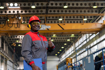 Cheerful African American Blue-Collar Worker Giving Thumbs Up In A Factory. Portrait Of Black Worker In Red Helmet, Safety Goggles, Hearing Protectors And Work Uniform. 