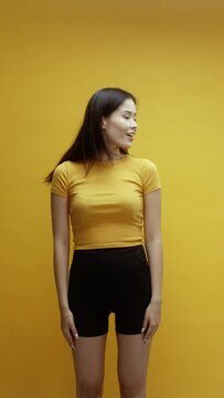 A young Asian woman being cute saying hellow and walking off camera, in front of a yellow background