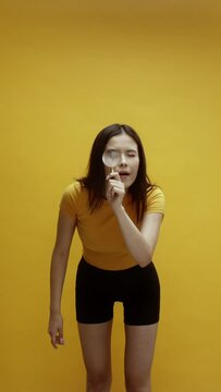 A young Asian woman searching with a magnifying glass, walking in front of a yellow background, wearing a yellow t-shirt