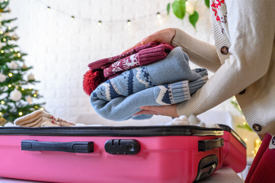 Christmas time. Woman putting knitted winter clothing in a suitcase in the room decorated christmas tree. Travel, holiday