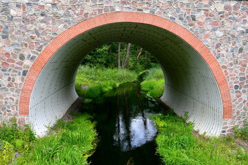 A view of a stone and brick bridge with an arch inside of it used to transport water with both banks of the water reservoir covered with grass, herbs, and other plants seen in Poland in summer