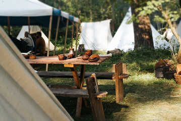 Tent medieval camp in the forest, a wooden table with pottery, people eating in the background in bokeh. Selective focus on ceramic kitchen utensils