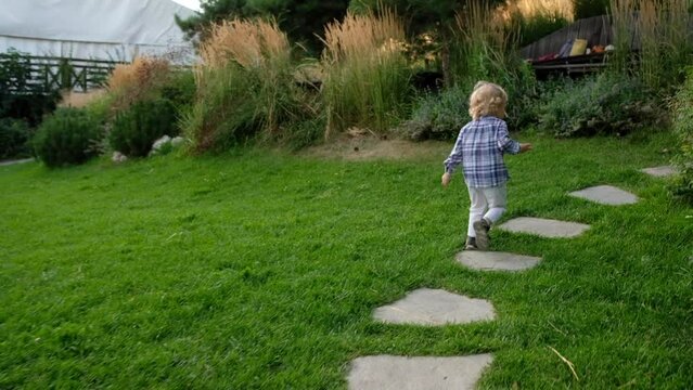 Little boy with white hair runs down path and falls in the park. Child stumble, fell and lies looking at camera, then gets up and goes on.