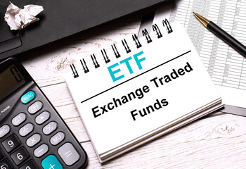 On a light-colored office table is a computer, a black calculator, a pen, and a notebook with the text ETF Exchange Traded Funds