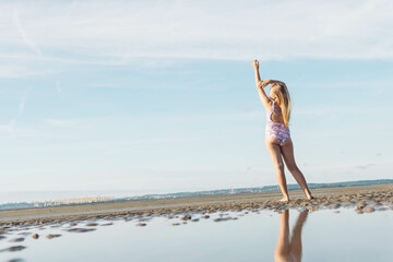 a teenage girl with blond long hair in a lilac swimsuit stands with her back to her full height on the ocean beach, in front of the photo there is water and on left there is place for an inscription