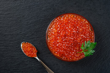 Delicious salmon caviar in a glass bowl over black slate background. Close-up of bowl and spoon...