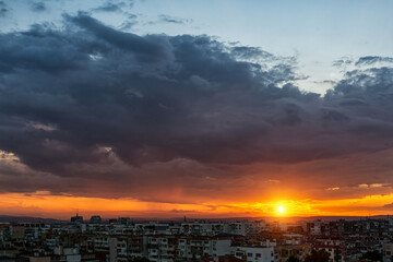 Sunset over multi-storey residential district. The setting sun low on the horizon while dark stormy cloud covering the blue sky. City residential area at sundown. Dramatic skyscape.