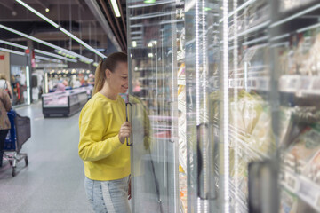 Fototapeta premium a woman of European appearance with blond hair tied in a ponytail in a yellow sweater and jeans, in a store in a refrigerated display case chooses products for the home. The concept of home food