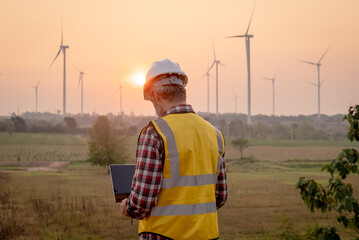 Portrait of engineer wearing yellow vest and white helmet using a computer laptop on site at wind turbines field or farm, Sustainable energy concept - obrazy, fototapety, plakaty