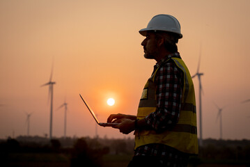 Portrait of engineer wearing yellow vest and white helmet using a computer laptop on site at wind turbines field or farm, Sustainable energy concept