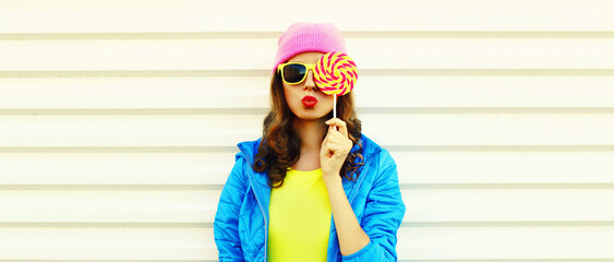 Portrait of stylish young woman blowing her lips with lollipop wearing colorful pink hat, blue jacket on white background