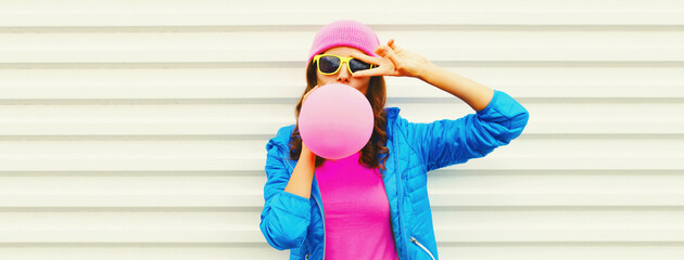 Fashionable portrait of stylish cool young woman inflating chewing gum or balloon wearing pink hat, blue jacket on white background