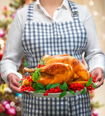 Woman holds golden roasted Christmas or Thanksgiving turkey garnished with red berries and sage leaves on festive light background
